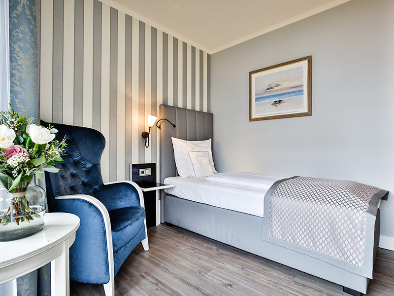 Helles, modernes Schlafzimmer mit einem Einzelbett, einem blauen Sessel, einer gestreiften Wand und einer Blumenvase auf einem Tisch.