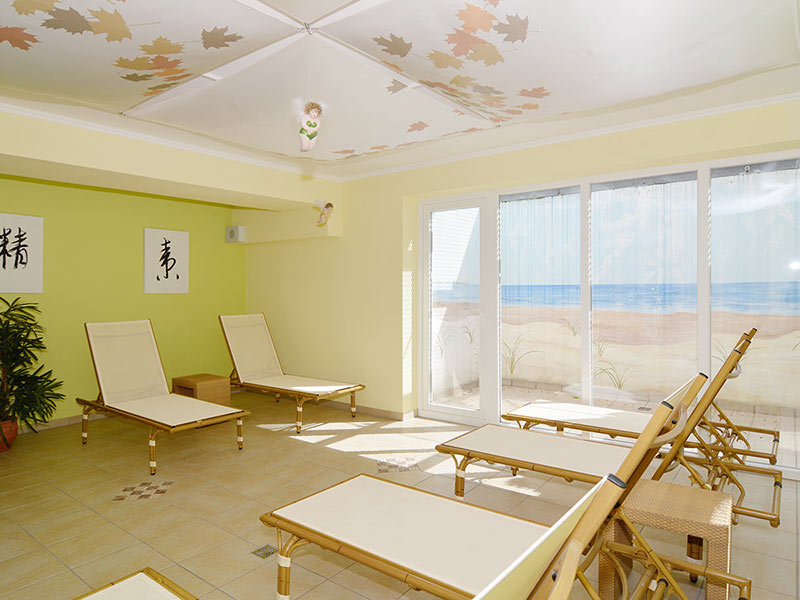 Heller Entspannungsraum mit Liegestühlen, Wandmalerei am Strand, Topfpflanzen und Sonnenlicht, das durch große Fenster einfällt.