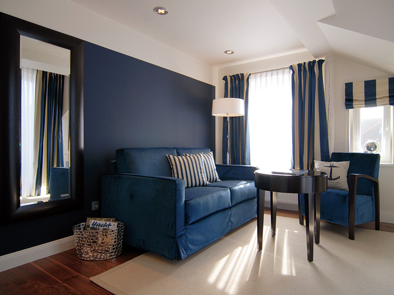Marinefarbenes Wohnzimmer mit blauem Sofa, gestreiften Kissen, rundem Tisch und großem Fenster mit gestreiften Vorhängen.