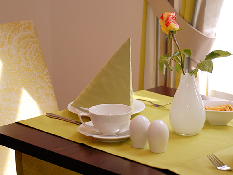 Ein gedeckter Tisch mit einer Teetasse, einer gefalteten Serviette, Salz- und Pfefferstreuern und einer Vase mit einer gelb-roten Rose.
